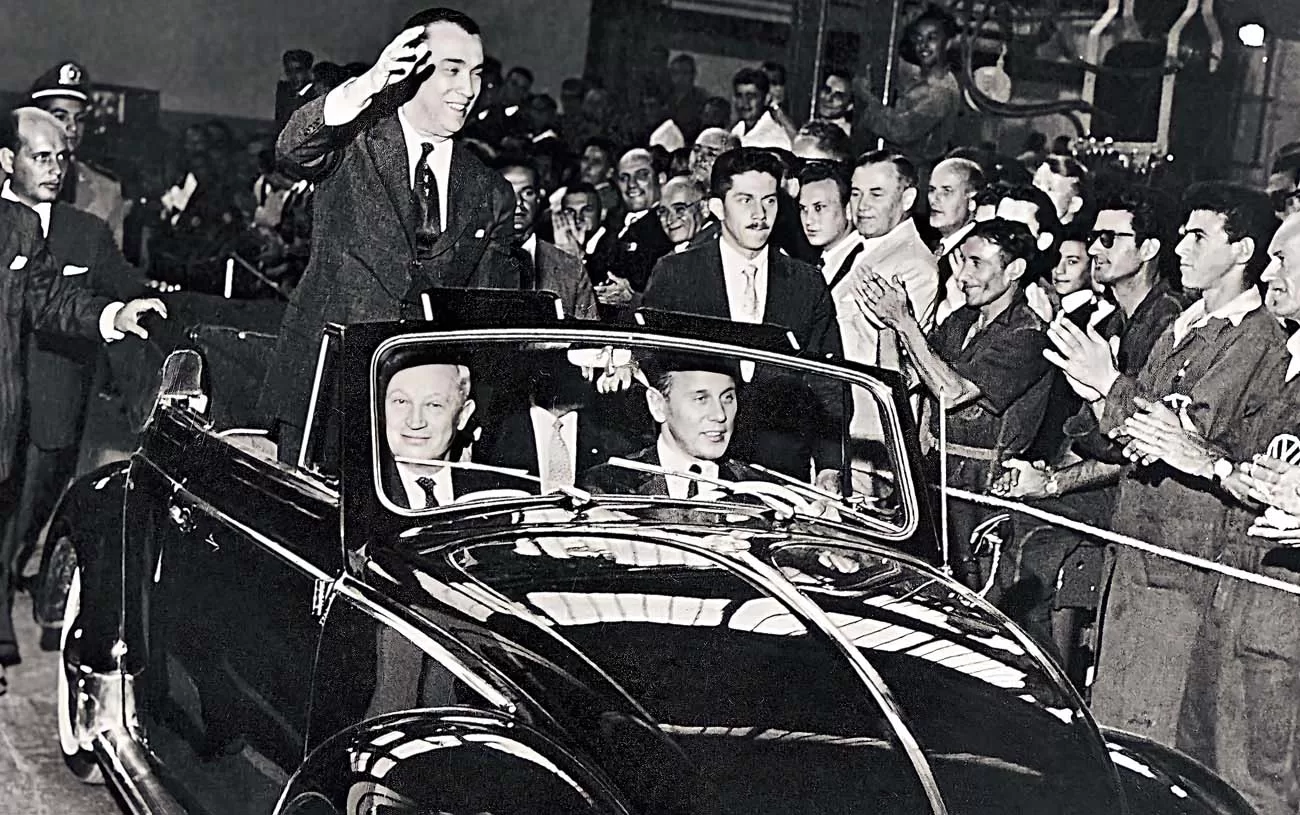 Juscelino Kubitschek acena a bordo de um Fusca conversível, dirigido por Wilhelm Shultz-Wenk, presidente da Volkswagen no Brasil, com Henrich Nordhoff, presidente do grupo Volkswagen, no banco de passageiros. (Imagem: Volkswagen/Divulgação)